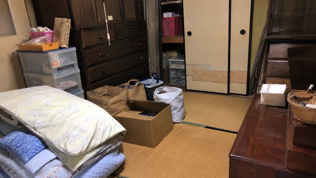 実家の片付け前、残った家具・布団・段ボール・古い木製家具を確認している和室のイメージ